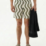 A front view of a model wearing The Shorts in Olive Curl Stripe by c'est normal, holding a black towel. The image is on a white background, highlighting the wavy pattern, elastic waist, and mid-thigh length of the versatile shorts.