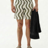 A front view of a model wearing The Shorts in Olive Curl Stripe by c'est normal, holding a black towel. The image is on a white background, highlighting the wavy pattern, elastic waist, and mid-thigh length of the versatile shorts.