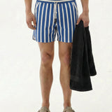 A model wearing The Shorts in Nautical Stripe, featuring a blue and white striped pattern, holding a black towel. The image showcases the shorts with their elastic waistband and drawstring detail, perfect for training or swimming by c'est normal.