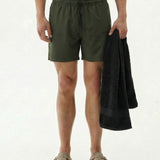 A front view of a model wearing The Shorts in khaki, holding a black towel, standing against a white background. The image highlights the elasticated waistband with a drawstring and the c'est normal logo.