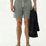 A front view of a model wearing The Shorts in a tile pattern by c'est normal, holding a black towel. The image showcases the shorts' elastic waistband, drawstring, and versatile design on a plain background.
