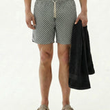 A front view of a model wearing The Shorts in a tile pattern by c'est normal, holding a black towel. The image showcases the shorts' elastic waistband, drawstring, and versatile design on a plain background.