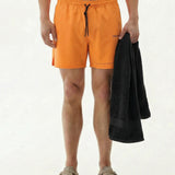 A model wearing The Shorts in Orange Peel on a white background, holding a black towel. The image shows the front view of the shorts featuring a black drawstring, notched hem, and small c'est normal text logo.
