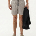 A shirtless model wearing The Shorts in Chalk by c'est normal, holding a black towel. The shorts feature an elasticated waistband with a black drawstring and a black c'est normal logo on the left leg.