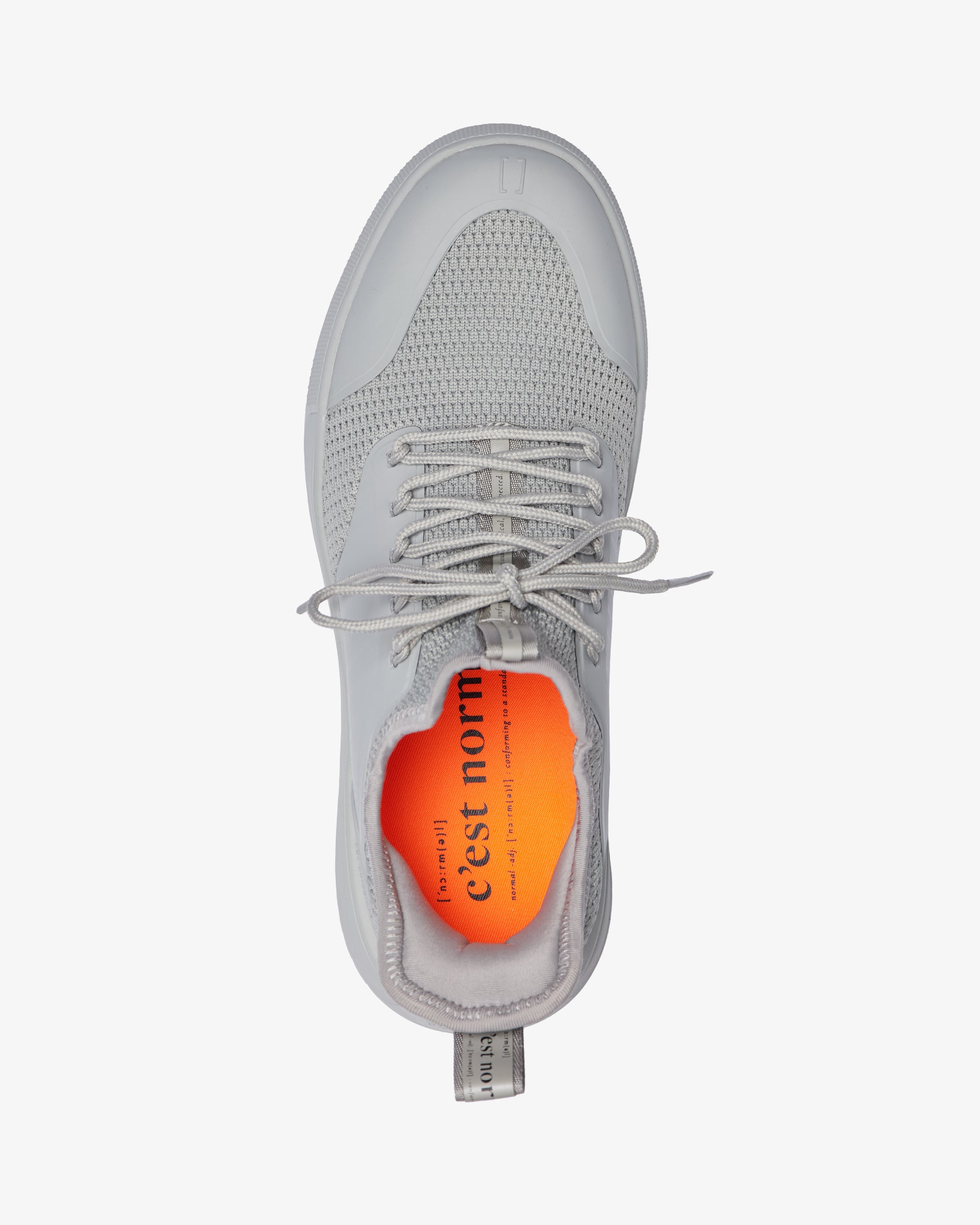 A top-down view of The Sneakers 2.0 in grey, laid flat on a white background. The image highlights the tone on tone mesh upper, custom lace loops, and the bright orange insole featuring the c'est normal logo.