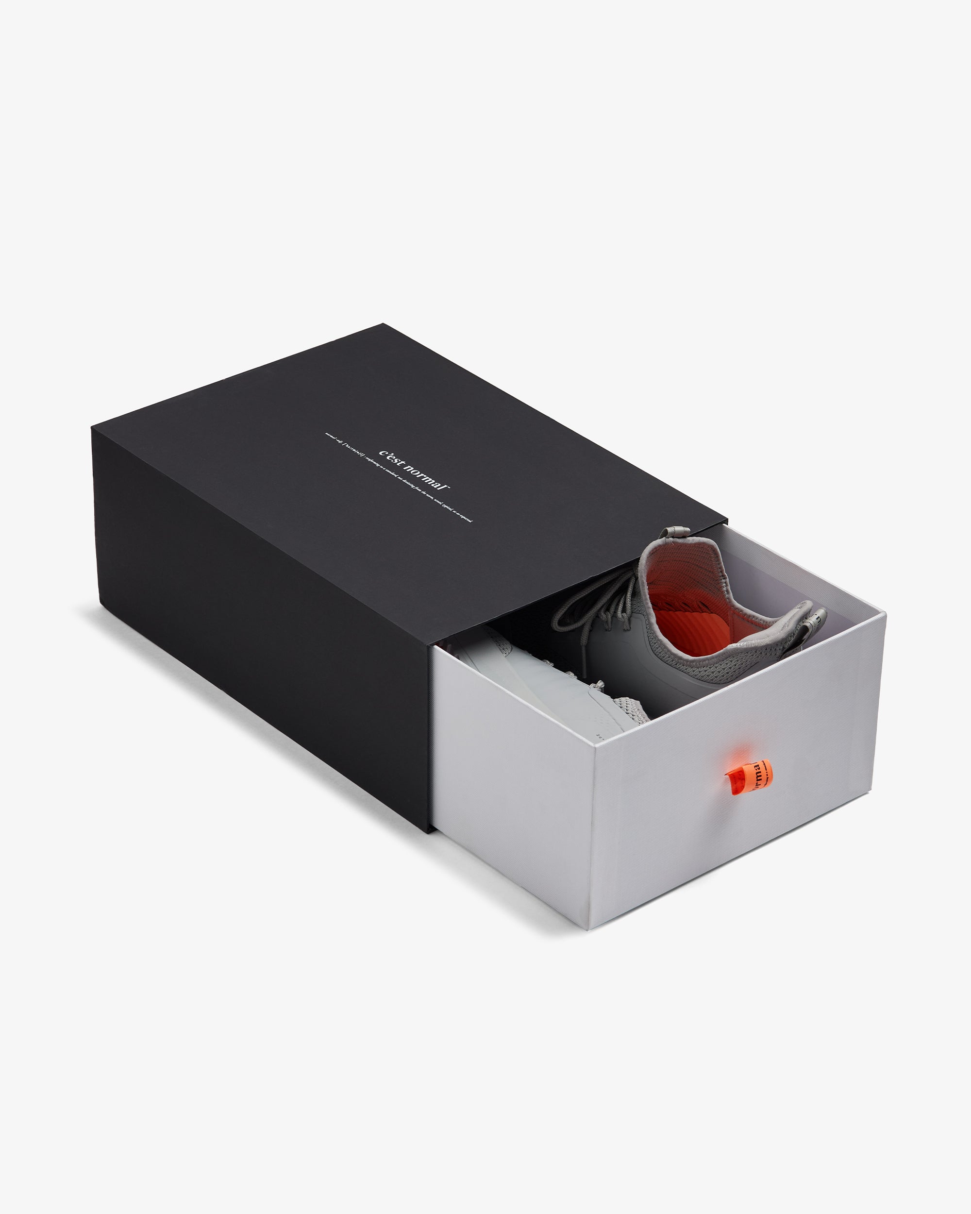 The Sneakers 2.0 in grey, partially pulled out of a black and white sliding shoebox on a white background. The black outer sleeve features the white c'est normal logo, and the inner white drawer has a bright orange pull tab.
